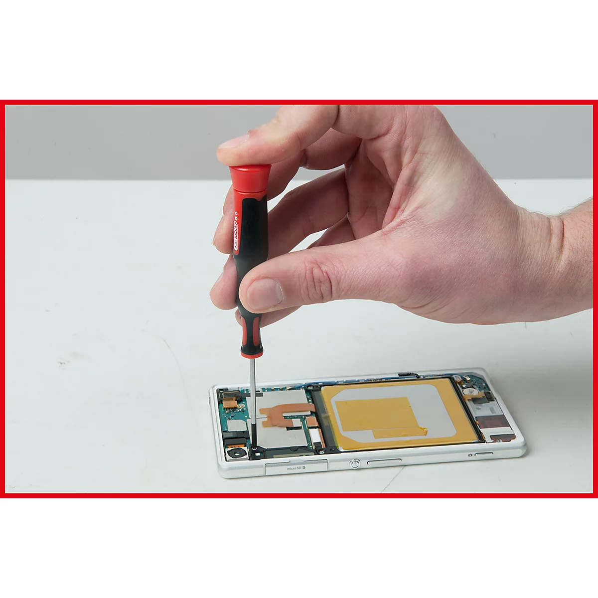 Hand hält Schraubendreher über einem geöffneten Smartphone. Auf hellgrauem Tisch.